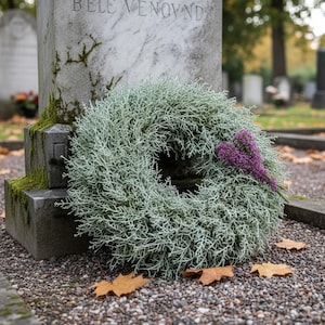 May include: A circular wreath made of silvery-green, fine-textured foliage with a small cluster of vibrant purple heather flowers attached to the right side, resting against a weathered gray stone monument with visible moss growth.