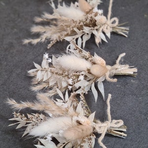 Könnte beinhalten: Drei kleine Sträuße getrockneter Blumen, die mit Schnur gebunden sind. Die Sträuße bestehen aus getrockneten Gräsern, weißem Pampasgras und kleinen weißen Blüten.