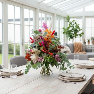 May include: A vibrant floral arrangement featuring a mix of dried and fresh botanical elements, including eucalyptus leaves, bright orange and pink plumes, red berries, and white pampas grass, centered on a light wood dining table with place settings.