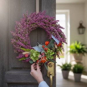 Könnte beinhalten: Ein runder Kranz aus leuchtend violetten Heidekrautblüten, akzentuiert mit grünen Hortensienblüten, leuchtend orangefarbenen Lampionblumen, kleinen roten Beeren und beigem Hasenschwanzgras, befestigt an einer dunklen Holztür mit Messinggriff.