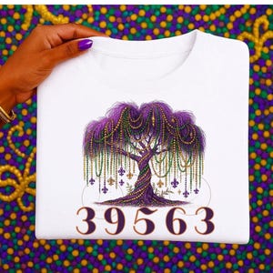 May include: White t-shirt featuring a purple tree adorned with green, gold, and purple beads, and fleur-de-lis symbols. The number "39563" is printed at the bottom. The shirt is held by a hand with purple nail polish.