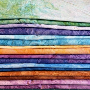May include: A stack of colorful fabric pieces in various shades of green, purple, blue, orange, and brown. The fabric appears to be batik or tie-dye, with unique patterns and textures. The fabrics are neatly folded and stacked, showcasing a variety of colors.