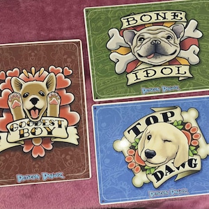 May include: Three metal signs with dog-themed designs. One sign features a smiling dog with the text "GOODEST BOY." Another shows a bulldog with "BONE IDOL." The third sign displays a winking dog with "TOP DAWG."