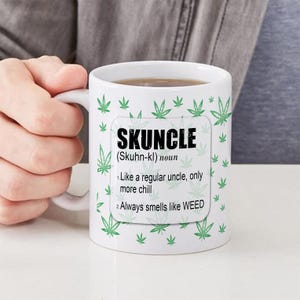 May include: White ceramic mug with the word "SKUNCLE" in bold black letters, with a definition. The mug is decorated with green cannabis leaves. The mug is filled with a brown liquid.