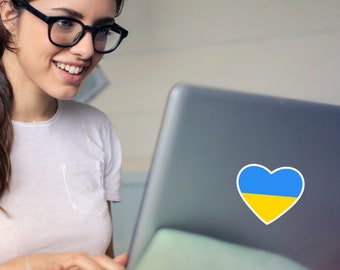Może przedstawiać: Kobieta w okularach pracuje na laptopie. Laptop ma naklejkę w kształcie serca w kolorach flagi ukraińskiej.