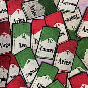 Puede incluir: Una colección de 12 parches para planchar que representan los signos del zodíaco. Cada parche tiene la forma de un paquete de cigarrillos con un fondo rojo y blanco o verde y blanco. Los parches están bordados con el símbolo del zodíaco y el nombre del signo.