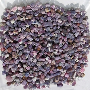 Könnte beinhalten: Eine große Sammlung kleiner, rauer, violetter und rosafarbener Edelsteine. Die Edelsteine sind in verschiedenen Formen und Größen vorhanden, und einige haben einen metallischen Glanz.