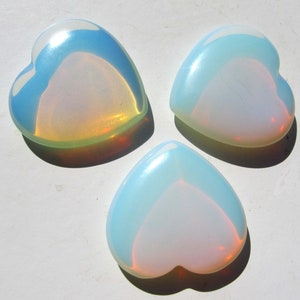 Könnte beinhalten: Drei herzförmige Opal-Steine, jeder mit einem anderen irisierenden Schimmer. Die Steine sind hellblau mit einem Hauch von Rosa und Gelb.
