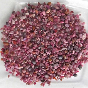 Könnte beinhalten: Eine Nahaufnahme einer Sammlung kleiner, rosafarbener, roter und violetter Edelsteine. Die Edelsteine sind in verschiedenen Formen und Größen und auf einer weißen Oberfläche verstreut.