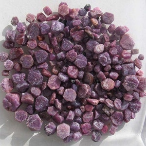 Könnte beinhalten: Eine Sammlung kleiner, rauer, violetter Edelsteine. Die Edelsteine sind in verschiedenen Violetttönen gehalten und haben einen leicht schillernden Glanz.