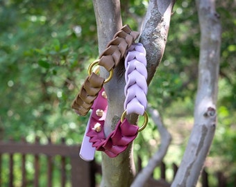 May include: Three braided leather dog collars in shades of brown, purple, and lavender hang from a tree branch. Each collar features a gold-colored ring and a matching leather tag. The purple collar has gold studs.