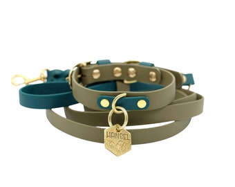 May include: Olive green dog collar and leash set with teal accents and gold-tone hardware. The collar has gold studs and a matching leash. A gold tag with the word "HANSEL" and a mountain design hangs from the collar.