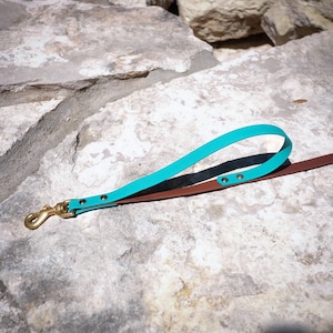 Op de afbeelding: Een turquoise en bruine leren hondenriem met een messing haak. De riem ligt op een lichtgrijs stenen oppervlak.
