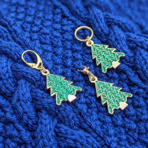 May include: Three gold-colored Christmas tree stitch markers with green and white glitter. The markers are on a blue knitted fabric.