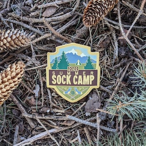May include: A brown and yellow embroidered patch with a mountain scene and the text "2022 Summer Sock Camp".