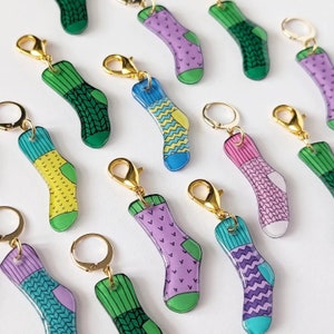 Pode incluir: Um conjunto de 12 marcadores de ponto de meia coloridos com fechos de lagosta dourados. Cada marcador apresenta um design de meia diferente com várias cores e padrões.
