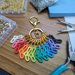 Op de afbeelding: Een gouden sleutelhanger met een ring van kleinere gouden ringen. Aan de ringen zitten 10 kleurrijke plastic sokkenvormige bedels in een regenboogpatroon.