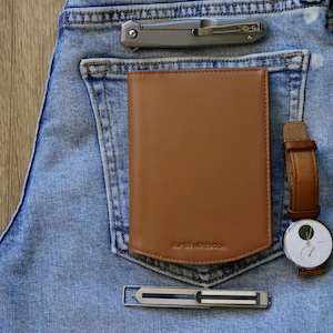 Puede incluir: Un cuaderno de cuero marrón claro con el texto "SUAVE NOTEBOOK" en el bolsillo trasero de unos vaqueros azules. También se ven una navaja de bolsillo plateada, un clip para billetes plateado y un reloj de pulsera con correa de cuero marrón.