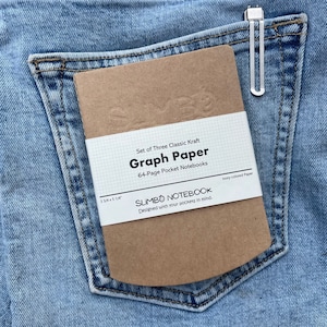 May include: A set of three brown kraft paper graph paper notebooks, each with 64 pages. The notebooks are 3 3/4 inches by 5 1/4 inches and are designed to fit in your pocket. The text on the front of the notebooks reads "Set of Three Classic Kraft Graph Paper 64-Page Pocket Notebooks SLIMBO NOTEBOOK Designed with your pockets in mind. Ivory-colored Paper."