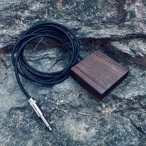 May include: A dark brown wooden square with a black cable attached to it. The cable has a silver 1/4 inch jack on the end.