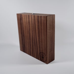 Puede incluir: Una caja de madera marrón oscuro con un pequeño pestillo de metal en la parte superior. La caja tiene un patrón de veta de madera y un pequeño logotipo en la parte delantera.