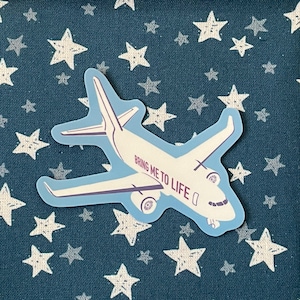 Pode incluir: Um autocolante de avião branco com detalhes azuis e o texto "BRING ME TO LIFE" num fundo azul marinho com estrelas brancas. O autocolante apresenta uma ilustração detalhada de um avião.