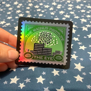 May include: Holographic sticker with a black border. The design features the text "THE MAGNUS INSTITUTE" and "THE EYE", a cluster of eyes above a cityscape, and a green and yellow colour scheme.