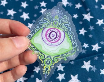 May include: A transparent sticker featuring a stylized eye design. The eye has a green iris with a purple and white pupil, surrounded by intricate, swirling patterns in green, yellow, and purple. The sticker is held against a blue fabric with white star patterns.