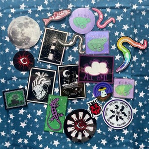 Puede incluir: Una colección de pegatinas coloridas con varios diseños, que incluyen un faro, un corazón, un barco, una rana, una nube, una luna, un gusano y una mano con dientes. Las pegatinas están sobre un fondo azul con estrellas blancas.