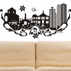 May include: Black and white silhouette of a city skyline with buildings, trees, and a car. The skyline is framed by a decorative floral design. The word "NIKOSIA" is written at the bottom of the design.