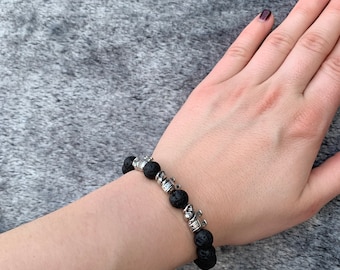 May include: A black lava rock bracelet with silver crown charms. The bracelet is on a person's wrist.
