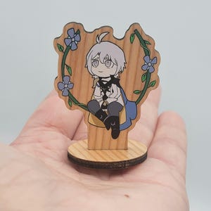May include: A wooden figurine featuring a cartoon character with white hair, wearing a white and blue outfit, and sitting within a floral wreath. The figurine is mounted on a round wooden base and a rectangular wooden stand.