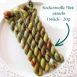 Könnte beinhalten: Vier Mini-Stränge aus meliertem Garn in Olivgrün, Salbei und Rostorange. Der Text Sockenwolle Mini einzeln 1 Stück = 20g ist sichtbar, mit einem Pfeil, der auf das Garn zeigt. Jeder Strang ist gedreht und besteht aus Wollfasern.