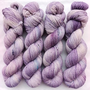 Könnte beinhalten: Vier handgefärbte Garnstränge, die nebeneinander angeordnet sind. Jeder Strang weist eine variierende Farbpalette aus sanften Lavendeltönen, tiefem Violett und Cremeweiß auf, mit feinen blauen und dunkelvioletten Sprenkeln im gesamten Garn.