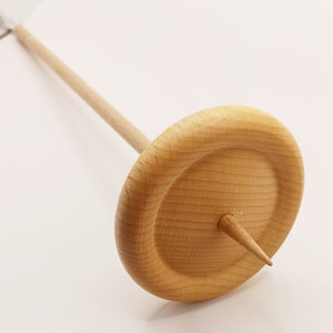 Könnte beinhalten: Eine Holzspindel zum Handspinnen von Garn. Sie hat einen langen, dünnen Schaft und einen kreisförmigen Wirtel. Der Wirtel hat eine spitze Spitze und ist aus hellfarbenem Holz gefertigt. Ein traditionelles Werkzeug für Textilhandwerk.