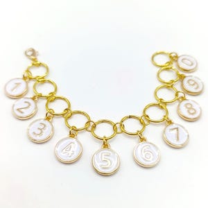 Könnte beinhalten: Ein goldfarbenes Bettelarmband mit zehn runden, weiß emaillierten Anhängern, die jeweils mit einer Ziffer von null bis neun geprägt sind. Die Anhänger sind an einer goldfarbenen Gliederkette mit einem Karabinerverschluss befestigt.