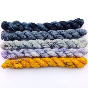 Könnte beinhalten: Fünf gedrehte Garnstränge, die horizontal angeordnet sind. Der oberste Strang ist dunkel anthrazit, gefolgt von einem mittleren Schieferblau, einem hellen Grau mit dunklen Sprenkeln, einem blassen Lavendel mit Sprenkeln und einem leuchtenden Goldgelb.