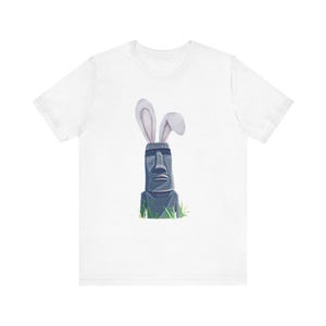 May include: White t-shirt with a gray stone head wearing bunny ears. The head is surrounded by green grass.
