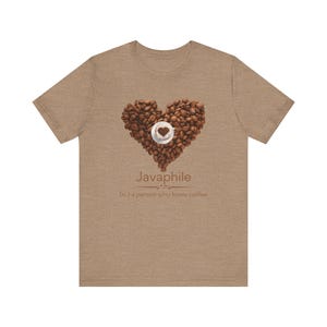 Pode incluir: Uma camiseta bege com um coração feito de grãos de café e uma xícara de café pequena. A palavra "Javaphile" é impressa abaixo do coração, com a definição "(n.) uma pessoa que ama café".