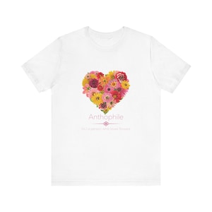 May include: White t-shirt featuring a heart-shaped floral design in shades of pink, yellow, and red. The word "Anthophile" is printed below the floral heart, with the definition "(n.) a person who loves flowers" underneath.