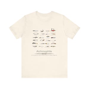 May include: A cream-coloured t-shirt displaying various knife illustrations. The word "Aichmophile" is printed below the knives, with the definition: "(n.) a person who loves knives or sharp objects."
