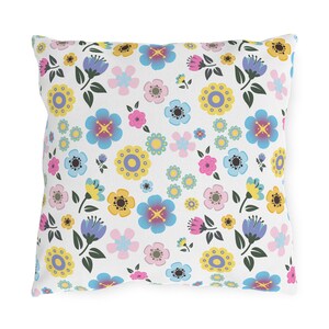 May include: Square pillow with a white background and a repeating floral pattern. The flowers are in shades of blue, yellow, pink, and purple, with green leaves. The pillow is a decorative home accent.