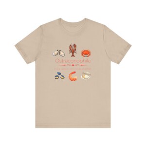 May include: A tan t-shirt with a graphic of various shellfish and the text "Ostraconophile (n.) a person who loves shellfish".