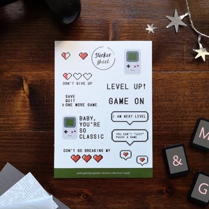 Könnte beinhalten: Eine Blatt mit Aufklebern mit einem Pixel-Spiele-Thema. Die Aufkleber zeigen Sätze wie "Gib nicht auf", "Spiel speichern Beenden", "Baby, du bist so klassisch", "Level Up", "Spiel an", "Ich bin auf dem nächsten Level", und "Du kannst nicht einfach das Spiel pausieren".
