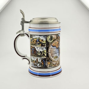 Può includere: Una boccale di birra in ceramica bianca con un coperchio in peltro. La boccale presenta una mappa del mondo dettagliata e varie scene di persone e animali. La boccale ha un bordo blu e rosso intorno alla parte superiore e inferiore.