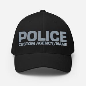 May include: Black baseball cap with the word "POLICE" in large, light gray letters across the front. Below the word "POLICE" is the text "CUSTOM AGENCY/NAME" in smaller, light gray letters. The cap has a curved brim.