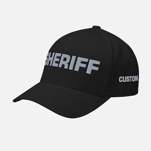 May include: Black baseball cap with the word "SHERIFF" in large, light gray letters across the front. The word "CUSTOM" is embroidered on the side. The cap has a curved brim and a structured fit.