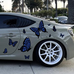 May include: A beige coupe car featuring multiple blue and black butterfly decals applied to the rear quarter panel and door. The vehicle is equipped with white multi-spoke alloy wheels and has dark tinted windows, showcasing a custom exterior design.