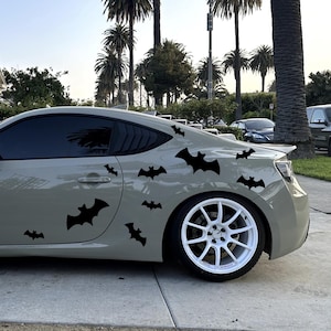 Può includere: Un'auto sportiva grigia con adesivi di pipistrelli neri sul lato. L'auto ha ruote bianche e vetri oscurati. L'immagine è stata scattata all'aperto con palme e una casa sullo sfondo.