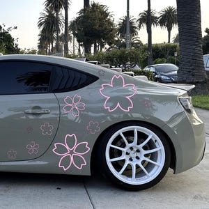 Puede incluir: Un coche deportivo gris con llantas blancas y calcomanías de flores de cerezo rosas. El coche está estacionado en una superficie pavimentada con palmeras al fondo. Las calcomanías son de varios tamaños y están colocadas en el lateral del coche.
